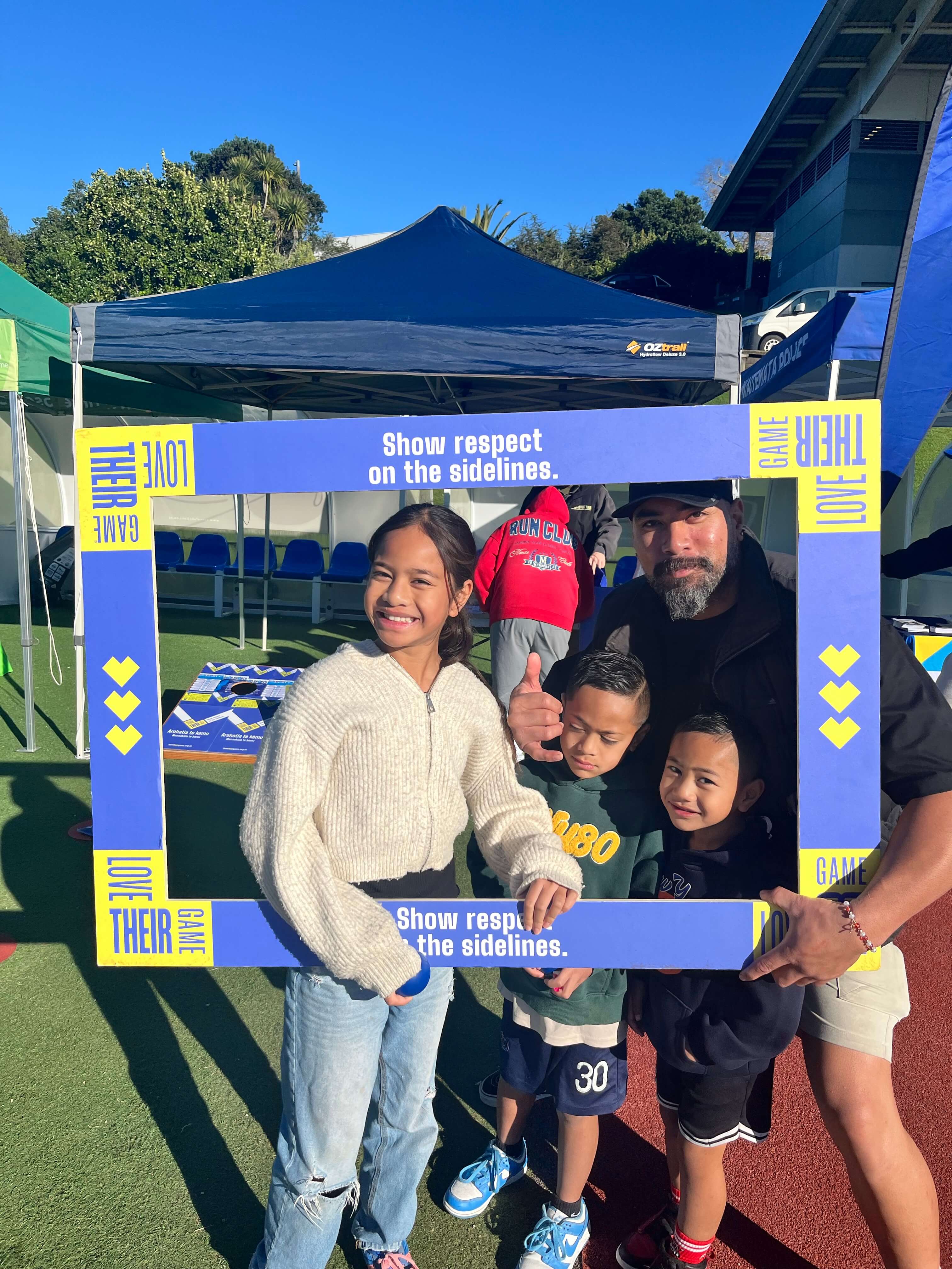 Whānau smiling inside Love Their Game frame at West Auckland Festival of Football